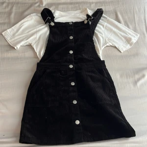 Svart manchesterklänning med hängslen från Lindex - Säljer en svart manchesterklänning från Lindex med justerbara hängslen och knappar framtill. Klänningen har fickor på sidorna och är i ett mjukt, strukturerat tyg. Perfekt för att styla med en t-shirt eller tröja under. Utmärkt att använda nu under de kommande sommardagarna. 🖤