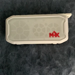 MAX bärbar högtalare - Vit bärbar högtalare från MAX med robust design och tydliga knappar för volym, play/pause och mode på ovansidan. Högtalaren har ett vitt metallgaller framtill med röd MAX-logga. Perfekt för att ta med sig musiken överallt. Kommer med en laddare