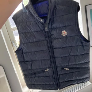 Mörkblå dunväst från Moncler - Snygg svart dunväst från Moncler med hög krage och dragkedja framtill. Västen har två fickor med dragkedja och Moncler-logga på bröstet. Perfekt för lager-på-lager under kyliga dagar. storlek 2