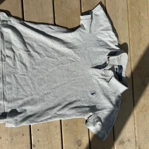 Grå pikétröja från Polo Ralph Lauren - Klassisk grå pikétröja från Polo Ralph Lauren med blå broderad logga på bröstet. Tröjan har korta ärmar, krage och knappar framtill. Slim fit-modell som passar perfekt till en avslappnad stil. Pris kan diskuteras. 