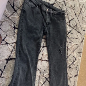 Svarta jeansbyxor med slitning - Säljer ett par svarta jeansbyxor med rak passform och slitning på ena benet. Klassisk femficksmodell och knappgylf. Perfekta för en avslappnad och trendig look.