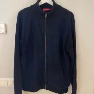 Svart stickad jacka med dragkedja från Redmond - Blå stickad jacka från Redmond med hög krage och hel dragkedja framtill. Jackan har ribbade muddar vid ärmslut och nederkant, vilket ger en snygg och bekväm passform. Perfekt för kyliga dagar.