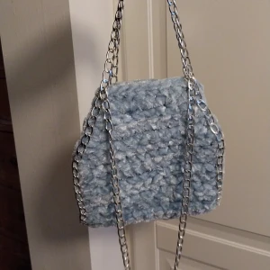 Ljusblå virkad handväska med kedja - Superfin ljusblå väska med virkad look och silvrig kedja. Perfekt för att lyfta en outfit med en unik accessoar. Väskan har en mjuk och fluffig känsla och är lätt att bära med sig.