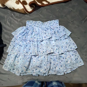 Blommig volangkjol i ljusblått - Supersöt ljusblå kjol med blommigt mönster och flera volanger. Kjolen har resår i midjan för extra komfort och en luftig känsla. Perfekt för dig som gillar romantisk och lekfull stil.