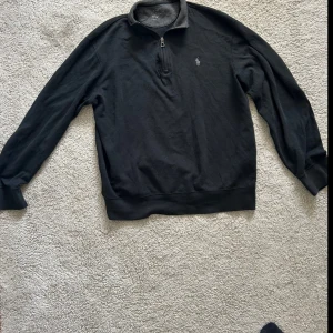 Svart tröja med dragkedja från Ralph Lauren - Klassisk svart långärmad tröja från Ralph Lauren med halv dragkedja och broderad logga på bröstet. Tröjan har ribbade muddar och krage, perfekt för en stilren och avslappnad look.