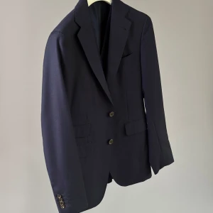 Mörkblå kostym från Rose & Born med väst - Säljer en klassisk mörkblå kostym från Rose & Born, tillverkad i Italien av Ermenegildo Zegna-tyg i superfine ull. Kavajen har två knappar, fickor med lock och matchande väst med fem knappar. Perfekt för bal eller fest.