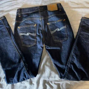 Mörkblå jeans från Nudie Jeans Co - Säljer ett par klassiska mörkblå jeans från Nudie Jeans Co med snygga kontrastsömmar och broderade bakfickor. Jeansen har fem fickor och normal passform. Perfekta för dig som gillar stilrena och tidlösa jeans. Modell thin finn 