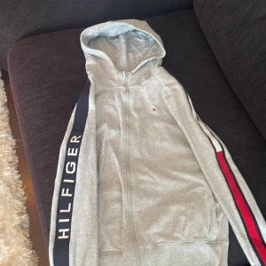 Grå hoodie från Tommy Hilfiger - Säljer en grå hoodie från Tommy Hilfiger med dragkedja och huva. Tröjan har långa ärmar med Hilfiger-tryck och röd, vit och blå rand längs ärmarna. Perfekt för en sportig och avslappnad stil.