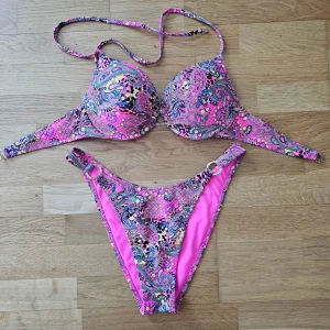 Färgglad paisleymönstrad bikini - Säljer en färgglad bikini med paisleymönster i pink, blått, svart och gult. Överdelen har vadderade kupor, bygel och knytband i nacken samt dekorativ ringdetalj framtill. Underdelen har högt skuren midja och matchande ringdetaljer på sidorna.Aldrig använd men tagit bort etiketten. Bikini passar till stl M och L 
