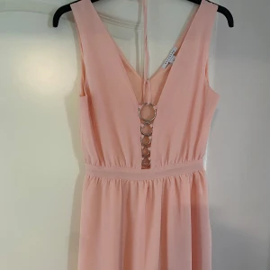 Ljusrosa ärmlös klänning med ringdetaljer - Söt ljusrosa klänning med djup v-ringning och dekorativa silverringar framtill. Klänningen är ärmlös, har markerad midja och lätt utställd kjol. Perfekt för sommarens tillfällen. Klänningen är enbart använd en gång.