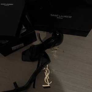 Svarta klackskor från Saint Laurent med YSL-klack - Säljer ett par svarta eleganta klackskor från Saint Laurent med öppen tå och ikonisk YSL-logga i guld som klack. Skorna har smal rem över tårna och runt vristen. Perfekta för dig som vill sticka ut med lyxig design. Säljer dessa eftersom jag köpte de bara för att använda dom till min bal! De kommer inte komma till användning längre.                      (ANVÄNT ENDAST EN GÅNG)!!! De är 100% äkta