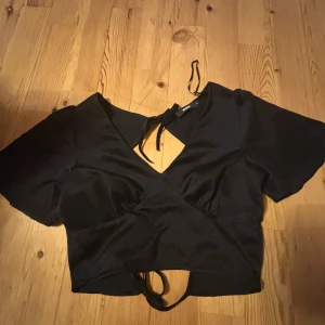 Svart croppad topp med knyt från BikBok - Säljer en svart blus från BikBok med v-ringning och knyt i ryggen. blusen har korta ärmar och är tillverkad i ett  kostym material. Perfekt för att skapa en trendig look.