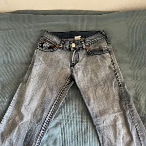 True Religion ljusblå jeans med kontrastsömmar - Säljer ett par ljusblå True Religion jeans med ikoniska kontrastsömmar och stora broderade bakfickor. Klassisk femficksmodell med knappgylf och tydlig logga på bakre linningen. Perfekta för dig som gillar streetwear och vill sticka ut.