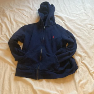 Mörkblå hoodie från Polo Ralph Lauren - Mörkblå hoodie från Polo Ralph Lauren med dragkedja och huva. Klassisk design med liten röd logga broderad på bröstet. Perfekt för en avslappnad stil och enkel att matcha med det mesta.