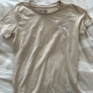 Beige t-shirt från Saint & Sinner - Säljer en beige t-shirt från Saint & Sinner med korta ärmar och rund halsringning. T-shirten har en vit broderad fågel på bröstet och en liten etikett nertill. Perfekt för en avslappnad stil.