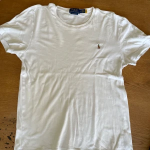 Vit t-shirt från Polo Ralph Lauren - Klassisk vit t-shirt från Polo Ralph Lauren med broderad logga på bröstet. Modellen är custom slim fit och har rund halsringning samt korta ärmar. Perfekt basic-plagg med stilren design.