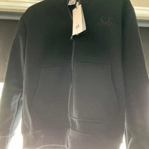 Svart hoodiejacka med glasögon från C.P. Company - Svart hoodiejacka från C.P. Company med dragkedja och ikoniska glasögon i huvan. Jackan har två stora fickor framtill och en stilren design med diskret logga på bröstet. Perfekt för dig som gillar streetwear och unika detaljer.