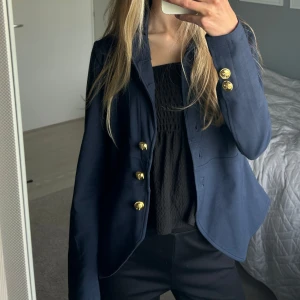 Mörkblå kavaj med guldknappar från House of lola o - Säljer en mörkblå kavaj med snygga, dekorativa guldknappar framtill och vid ärmsluten. Kavajen har en klassisk passform och är tillverkad i ett slätt tyg som ger en stilren look. Perfekt för att lyfta vilken outfit som helst. Saknas en knapp på höger armen men det medkommer en likadan extra knapp som går att sy fast. I övrigt ser den ut som ny. Säljer pga att den sällan kommer till användning. 