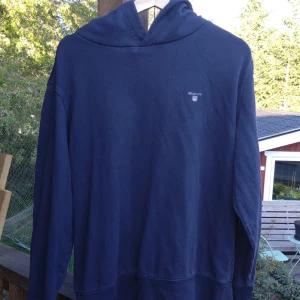 Mörkblå hoodie från GANT - Säljer en mörkblå hoodie från GANT med liten broderad logga på bröstet. Klassisk modell med huva och lång ärm, perfekt för en avslappnad stil.