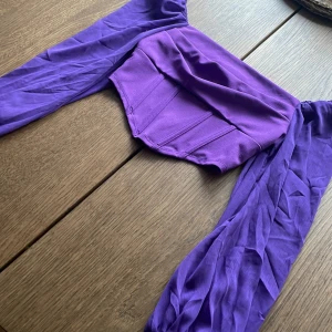 Lila croppad topp med långa ärmar från SHEIN - Säljer en lila croppad topp från SHEIN med långa, transparenta ärmar och dragkedja i ryggen. Toppen har en figurnära passform och är tillverkad i ett glansigt syntetmaterial. Perfekt för att sticka ut med en färgstark look! (Aldrig använd)