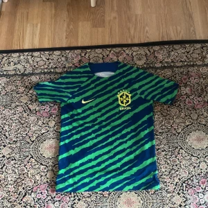 Brasilien fotbollströja Nike Dri-Fit - Säljer en Brasilien fotbollströja från Nike i Dri-Fit material. Tröjan är blå med gröna diagonala ränder och har gula detaljer, inklusive Brasil-logga och Nike-märke. Kortärmad och perfekt för fotbollsträning eller support.