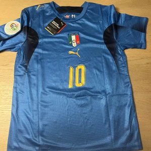 Italien fotbollströja Puma Totti #10 - Blå officiell Italien fotbollströja från Puma med Totti och nummer 10 i gult. Kortärmad modell med Italiens landslagsmärke på bröstet och Puma-logga. Perfekt för fans av italienska landslaget och Totti.