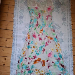 Vit långklänning med fjärilsmönster - Superfin långklänning i vit mesh med färgglada fjärilar och blommor i rosa, orange, blått och grönt. Klänningen har smala axelband och en figurnära passform upptill. Perfekt för dig som vill sticka ut med ett unikt mönster!
