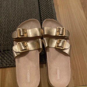 Guldiga sandaler med plattform och spännen - Säljer ett par snygga sandaler i guld med två breda remmar och stora guldfärgade spännen. Skorna har en vit plattformsula med korkdetalj på sidan och räfflad undersida. Perfekta för att lyfta din sommarstil!