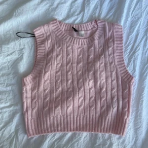 Ljusrosa stickad väst från H&M Divided - Säljer en ljusrosa stickad väst från H&M Divided i storlek XS. Västen har ett klassiskt kabelstickat mönster framtill och bred ribbad kant nedtill och vid ärmöppningarna. Perfekt att styla över en skjorta eller t-shirt.