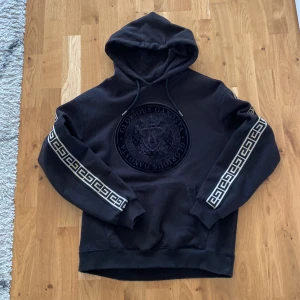 Svart hoodie från Glorious Gangsta - Säljer en svart hoodie från Glorious Gangsta med stort broderat emblem på bröstet och mönstrade band längs ärmarna. Tröjan har huva med dragsko och klassisk känguruficka framtill. Perfekt för en streetwear-look.