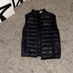 Svart dunväst från Emporio Armani EA7 - Snygg svart dunväst från Emporio Armani EA7 med guldfärgad logga på bröstet. Storlek M funkar bra till S med! Ny pris 1999kr Väldigt bra skick 10/10 använd bara 3 gånger! Västen har quiltad design och dragkedja framtill. Perfekt för lager-på-lager och enkel att ta med tack vare medföljande påse. 