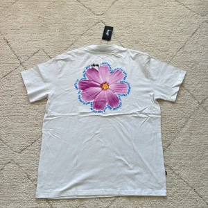 Vit t-shirt med blommotiv från Stüssy - Vit t-shirt från Stüssy med stor lila blomma tryckt på ryggen och mindre blått blommotiv på bröstet. Rund halsringning och normal passform. Perfekt för dig som gillar streetwear och snygga prints.