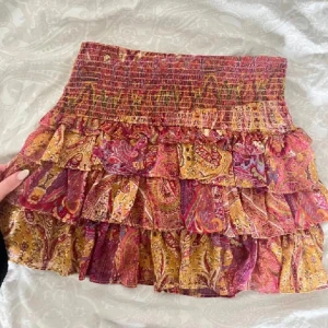 Mönstrad volangkjol i gult och vinrött - Supersöt kort volangkjol med paisleymönster i vinrött, gult och inslag av lila. Kjolen har flera lager volanger och smock i midjan för en bekväm passform. Perfekt för dig som gillar boho-stil och färg!