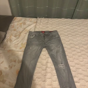 Guess jeans  - Säljer ett par ljusgrå jeans från Guess med slitningar på ena benet och klassisk femficksdesign. Byxorna har normal passform och raka ben. Perfekta för dig som gillar en avslappnad men ändå stilren look. Den är storlek XS men passar både S och M 