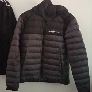 Svart dunjacka från Sail Racing - Säljer en svart dunjacka från Sail Racing med huva och dragkedja. Jackan har två sidofickor med dragkedja och logga på bröstet samt på ryggen. Perfekt för kyliga dagar och riktigt snygg passform.