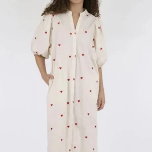 Vit klänning med röda hjärtan från Neo Noir - Supersöt vit långklänning från Neo Noir med broderade röda hjärtan. Klänningen har puffiga trekvartsärmar och en v-ringad halsringning. Perfekt för dig som vill sticka ut med en lekfull och romantisk stil.