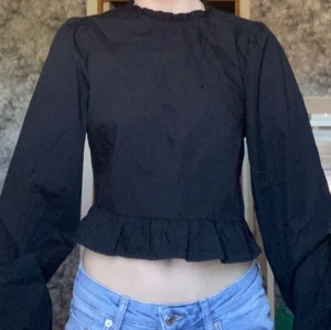 Svart blus med volang och dragkedja - Säljer en svart blus med långa puffärmar, volangdetalj vid nederkanten och liten volang vid halsen. Blusen har dragkedja i sidan och knytning i ryggen. Aldrig använd🩷 Perfekt för en stilren look!