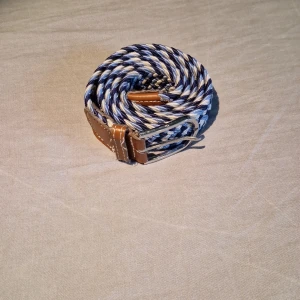 Blåvitt flätat bälte med läderdetaljer - Säljer ett snyggt flätat bälte i blått, vitt och beige med bruna läderdetaljer vid spännet. Bältet har ett klassiskt silverfärgat spänne och passar perfekt till jeans eller chinos. Ger en stilren och fräsch look till outfiten.