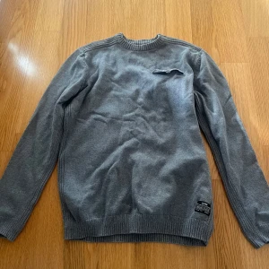 Grå stickad tröja från Jack & Jones - Säljer en stilren grå stickad tröja från Jack & Jones Core. Tröjan har rund halsringning, lång ärm och en liten fickdetalj med knapp på bröstet. Perfekt för lager-på-lager och enkel att matcha med jeans eller chinos.