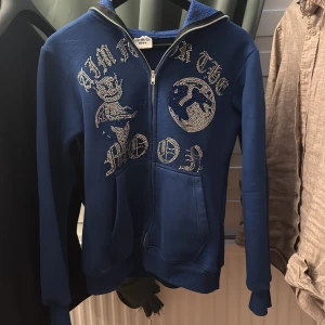 Blå hoodie med strassdetaljer och dragkedja - Säljer en blå hoodie med dragkedja och huva. Framsidan har stora strassdetaljer med text och motiv. Tröjan har långa ärmar och ribbade muddar. Perfekt för dig som gillar statement-plagg! Märket är aim for the moon 