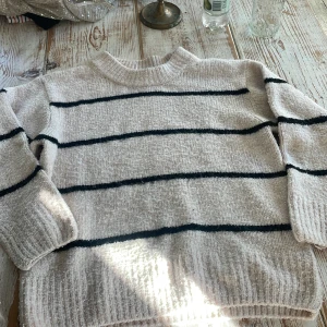 Beige stickad tröja med svarta ränder från Gina Tricot - Mysig beige stickad tröja från Gina Tricot med svarta horisontella ränder. Tröjan har rund halsringning och långa ärmar. Perfekt för kyligare dagar och enkel att matcha med jeans eller kjol.