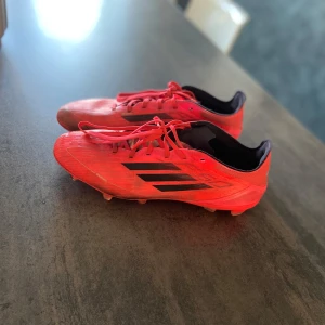 Röda Adidas fotbollsskor med svarta detaljer - Säljer ett par röda Adidas fotbollsskor med svarta ränder på sidan. Skorna har snörning och är designade för spel på gräs med dobbar undertill. Insidan är svart och ovandelen är i syntetmaterial. Skorna är PRO vilket är det näst dyraste alternativet inga defekter bara lite missfärgning 