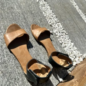 Bruna sandaler med svart häl - Snygga sandaler i brunt med öppen tå och svart hälparti i glansigt material. Platt sula och enkel design som passar perfekt till varma dagar. Bekväma och stilrena för sommaren. Lite slitna i hälen 