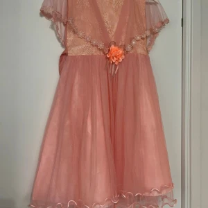 Rosa klänning med spets och blomdetalj - Söt rosa klänning med v-ringning, spetsdetaljer och en dekorativ blomma i midjan. Klänningen har korta ärmar i transparent tyg och volangkant nertill. Perfekt för speciella tillfällen. För barn som är 11-12 år gammal.