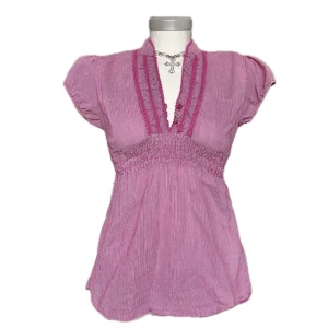 Rosa randig blus med v-ringning - Säljer en söt rosa blus med vita ränder och v-ringning. Blusen har korta puffärmar, smock i midjan och dekorativa knappar framtill. Finns några enstaka lösa trådar, vilket syns i bilderna. Lätt att klippa bort! 💓