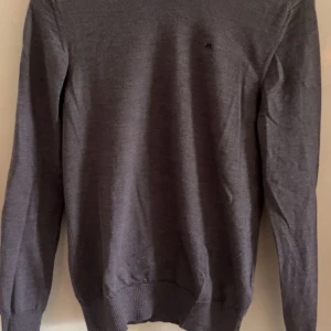 Grå stickad tröja från J.Lindeberg - Säljer en stilren grå stickad tröja från J.Lindeberg. Tröjan har rund halsringning och långa ärmar, perfekt för lager-på-lager. Diskret logga på bröstet.