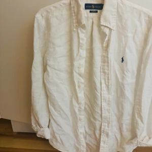 Vit linneskjorta från Ralph Lauren - Klassisk vit skjorta i linne från Ralph Lauren med blå broderad logga på bröstet. Skjortan har lång ärm, knappar framtill och en avslappnad passform. Perfekt för en stilren och fräsch look.