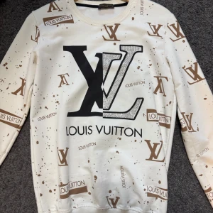 Vit Louis Vuitton sweatshirt med logotypmönster - Vit sweatshirt från Louis Vuitton med bruna LV-loggor och text tryckt över hela tröjan. Stor svart och brun LV-logga framtill. Rund halsringning och långa ärmar. Mjuk och bekväm, perfekt för en trendig streetstyle-look.