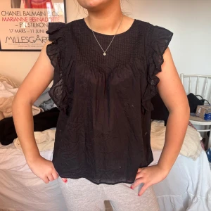 Svart volangblus från Indiska - Säljer en svart blus från Indiska med volangdetaljer längs axlarna och ärmlös design. Blusen har rund halsringning och ett lätt, luftigt tyg som passar perfekt till många olika outfits.