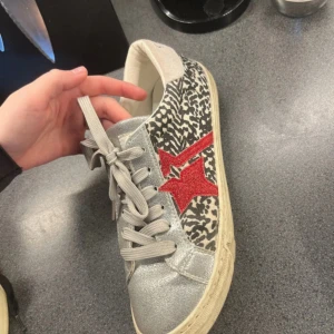Kaporal sneakers med zebramönster och röd stjärndetalj - Säljer ett par unika Kaporal sneakers med zebramönster på sidorna, silverfärgade detaljer och en röd stjärna på utsidan. Skorna har grå snörning och vit sula. 